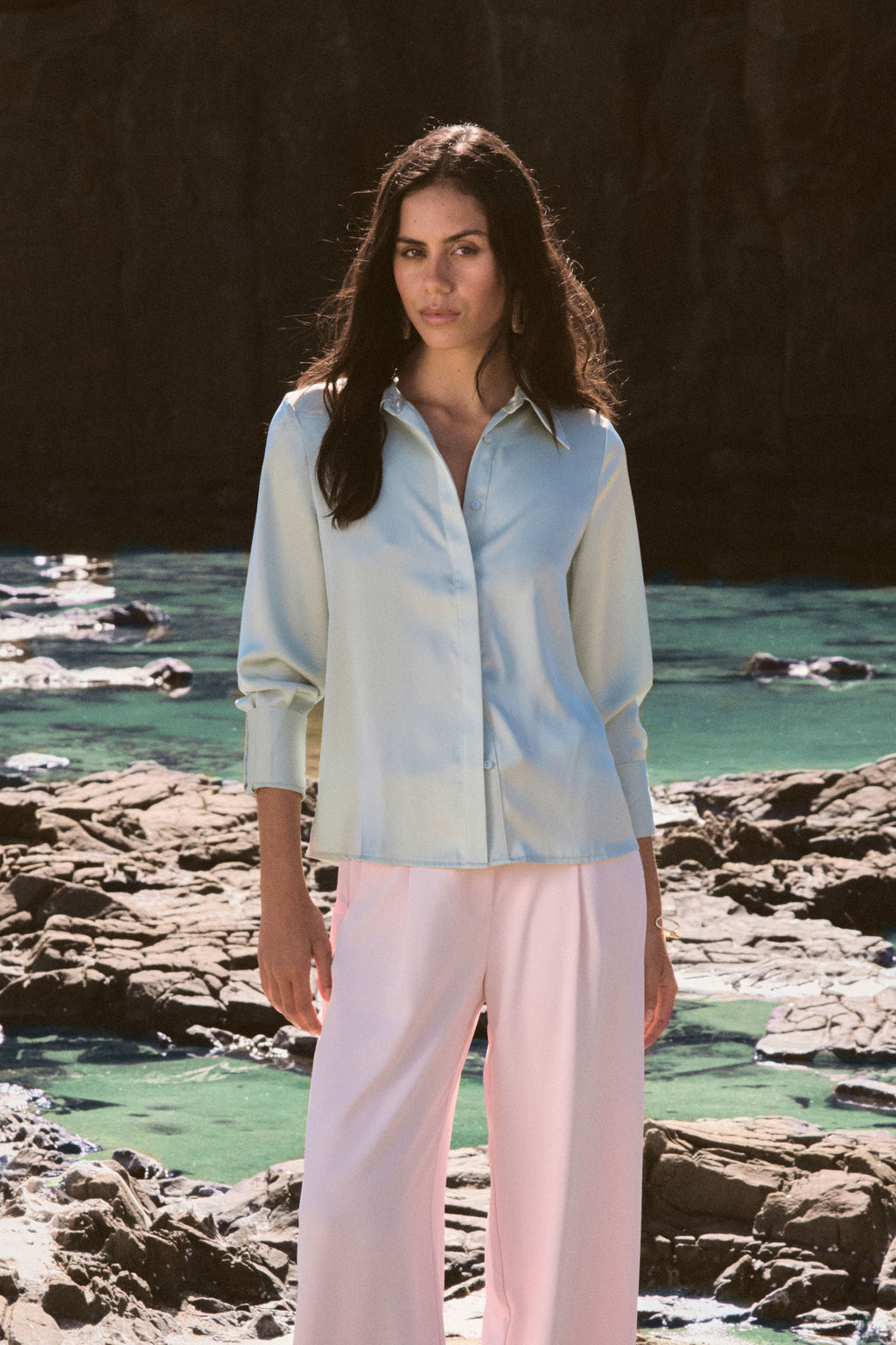 Mae Satin Shirt in mint worn front on with pink wide-leg trousers, coastal rocks behind